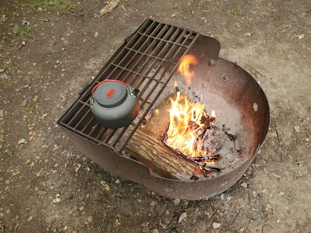 Un petit barbecue rouillé avec des bûches en flamme à l'intérieur, sur une mini grille est posé une bouilloire, le sol est en terre.