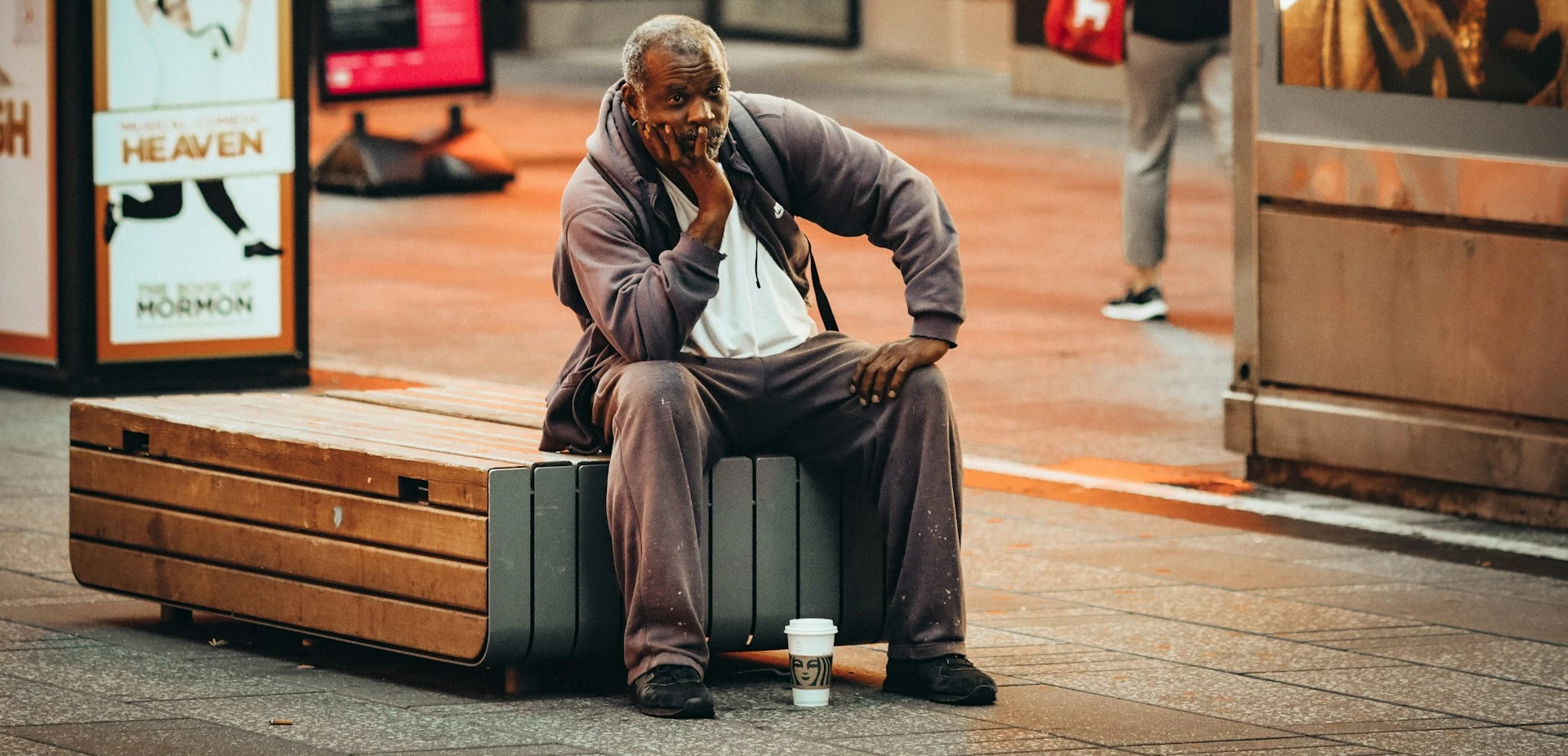 Homme assis en extérieur sur un banc, l'air désabusé, une main sous le menton, un café à ses pieds.